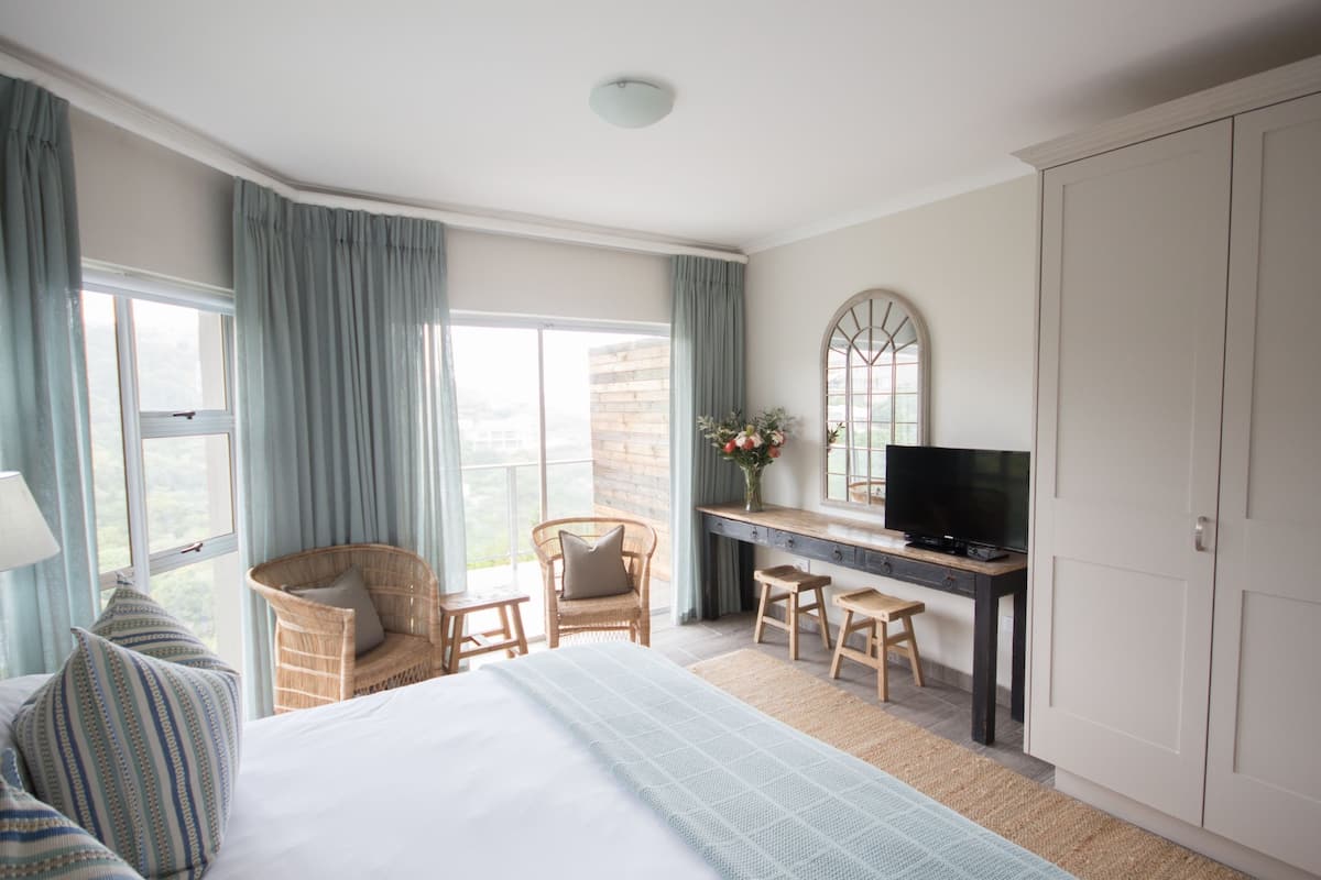 Hotelkamer met een bed, een bureau met televisie, twee stoelen, een groot raam met gordijnen en een garderobe.