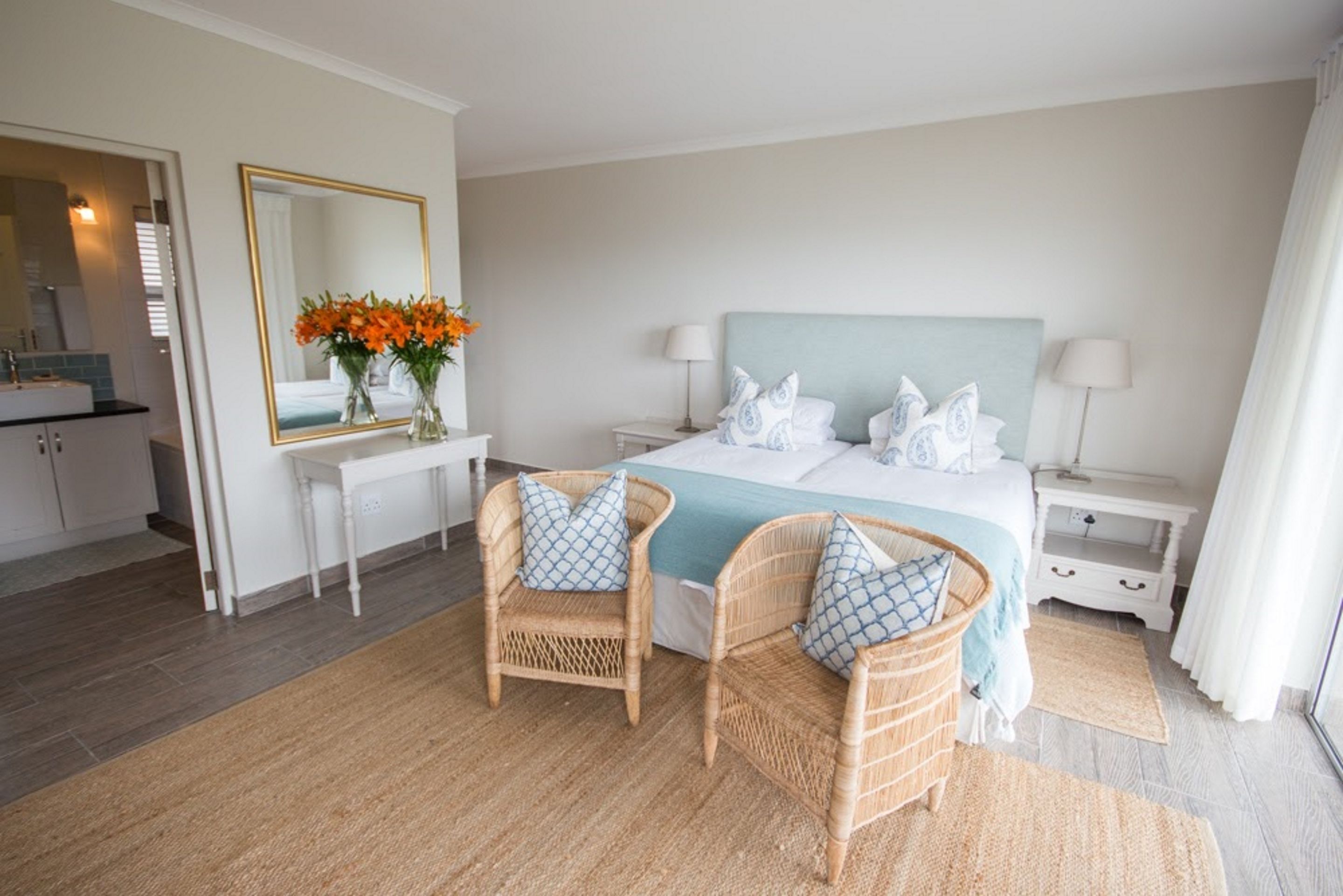 Quarto de hotel com uma cama, duas cadeiras de vime, uma mesinha de cabeceira com um vaso de flores, um espelho e uma mesa de apoio com um abajur.