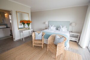 Una habitación de hotel con cama, dos sillas de mimbre, una mesita de noche con un florero y un espejo, junto a una mesita con lámpara.
