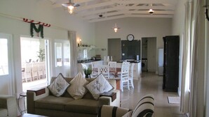 Amplia sala de estar con sofá y sillones, una mesa de centro de madera y un comedor con mesa y sillas. Se observa una cocina con fregadero y estufa al fondo.