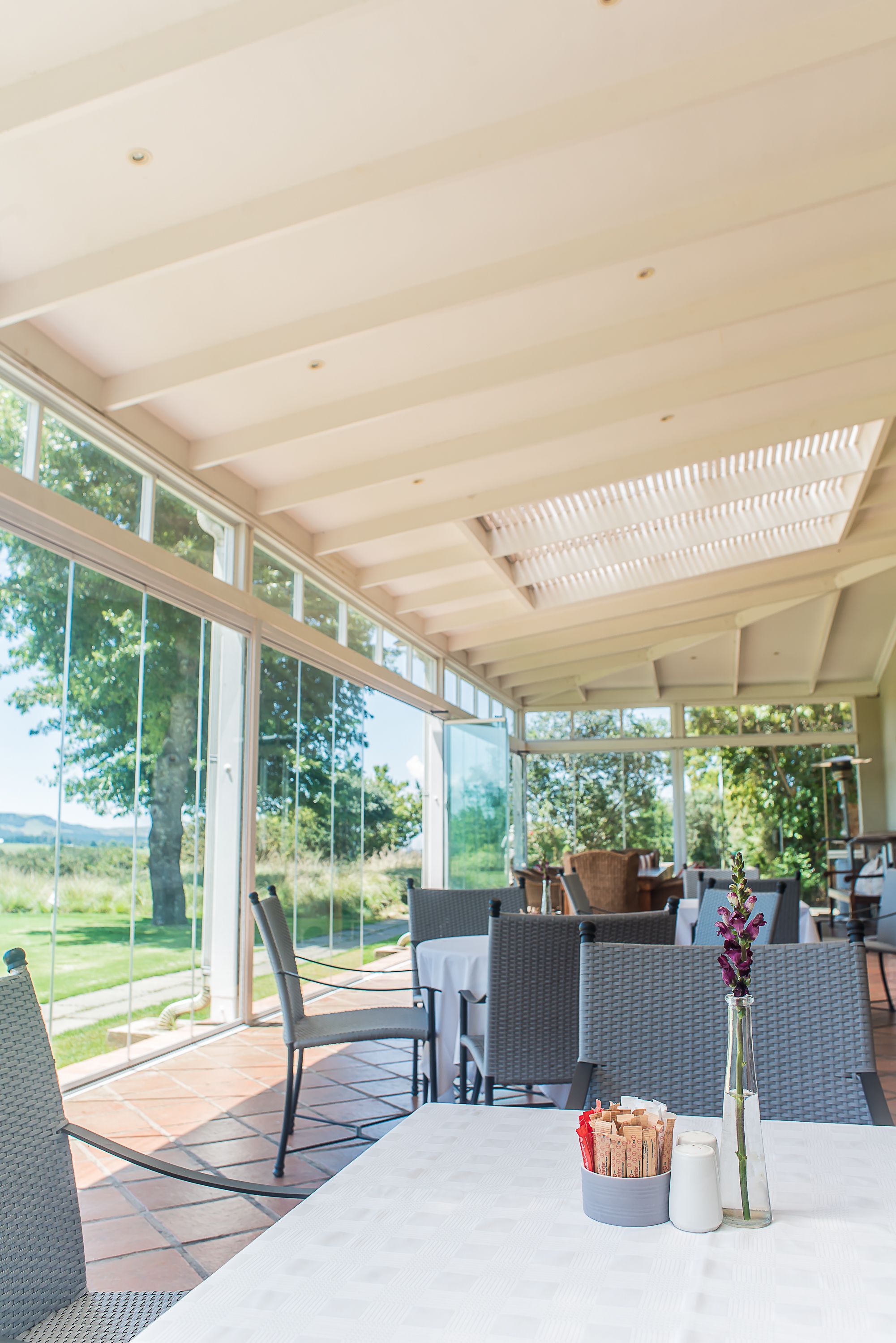 Un patio cubierto con mesas y sillas, ventanas amplias y vistas al exterior.