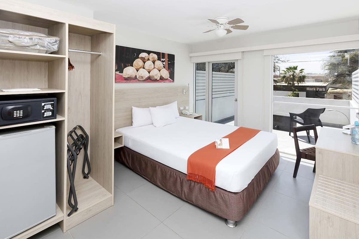 A hotel room with a bed, a desk, a chair, a wardrobe, and a balcony with a view of a park.