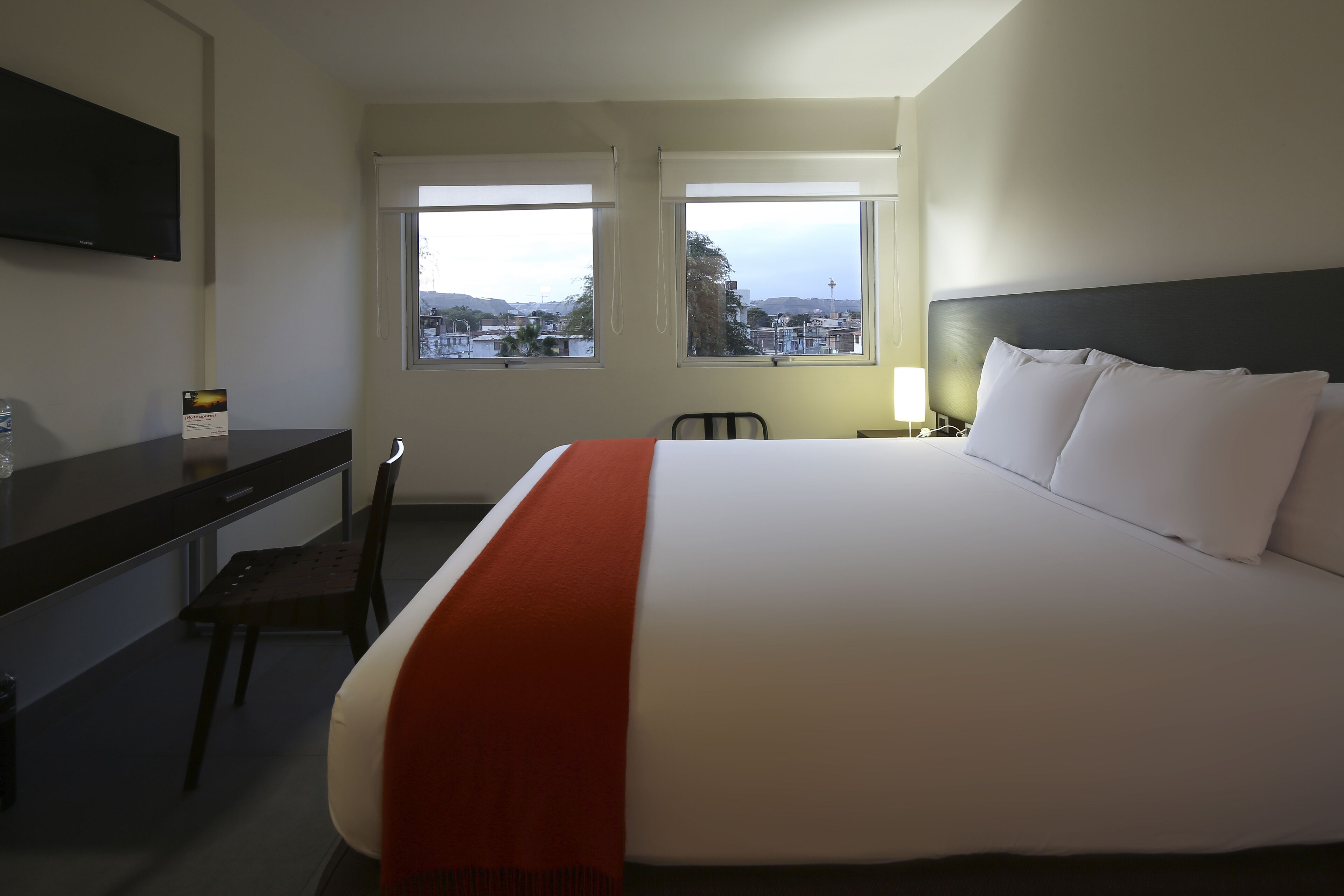 Une chambre d’hôtel avec un grand lit, un bureau et deux fenêtres donnant sur la ville.