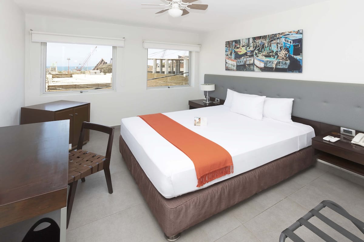 A hotel room with a large bed, a desk, a chair, a nightstand with a lamp, and a painting on the wall.