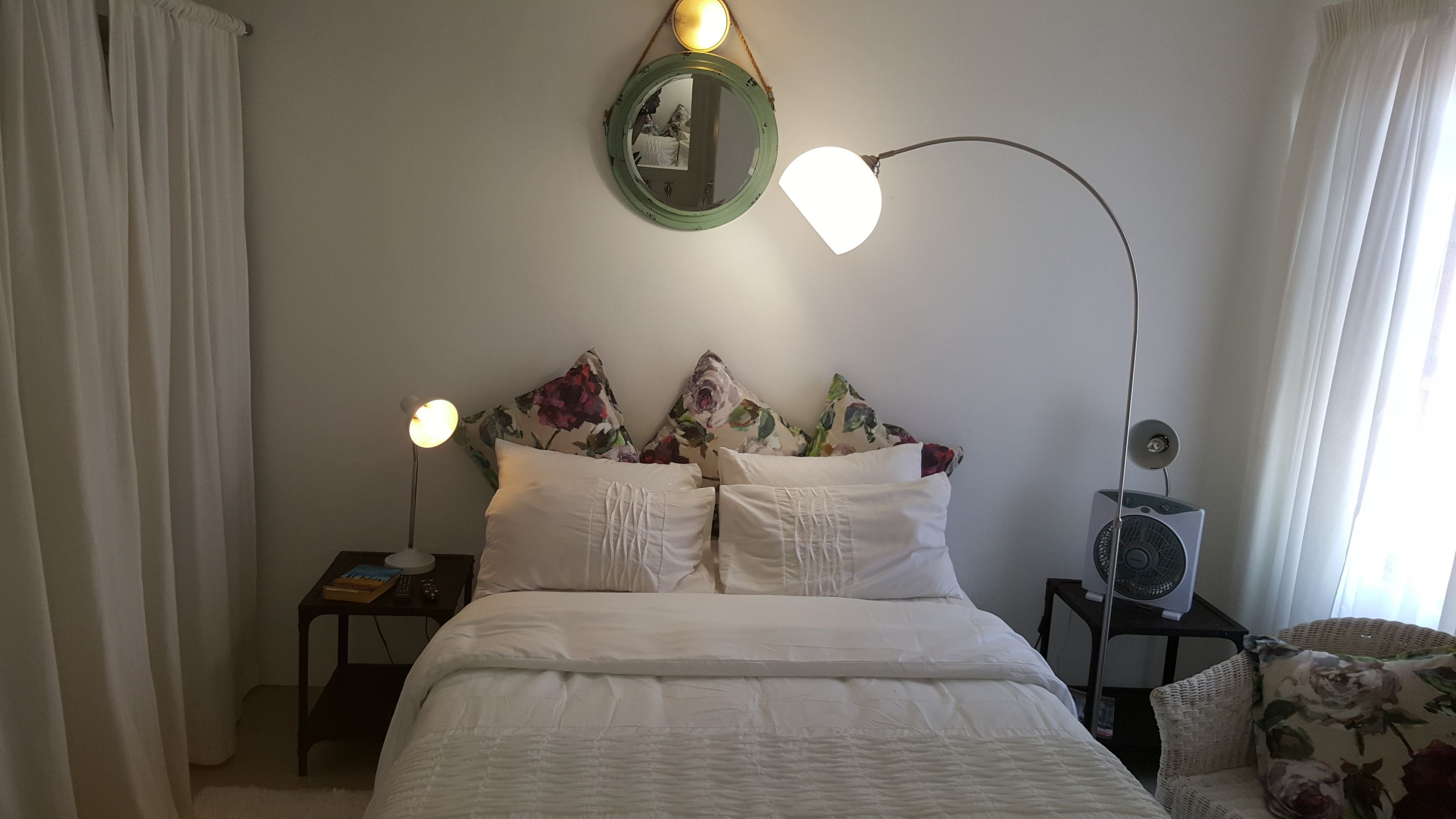 A bedroom with a bed, two bedside lamps, a mirror, a fan, and a window with curtains.