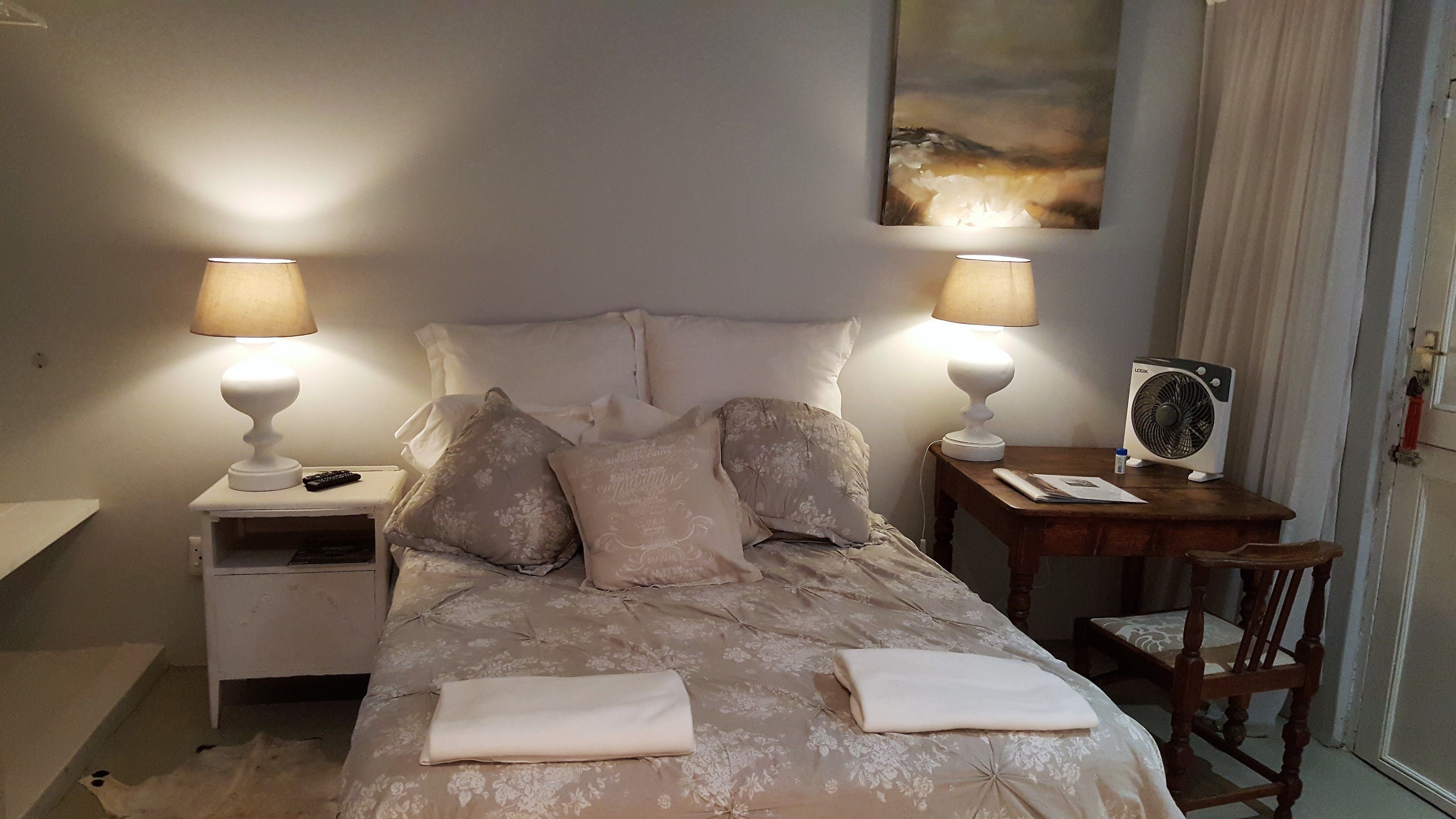 A neatly made bed with a floral bedspread, two bedside lamps, a nightstand with a remote control, a wooden nightstand with a fan, and a chair.
