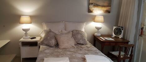 A neatly made bed with a floral bedspread, two bedside lamps, a nightstand with a remote control, a wooden nightstand with a fan, and a chair.