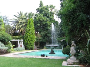 Un jardín con una fuente, una piscina y abundante vegetación.