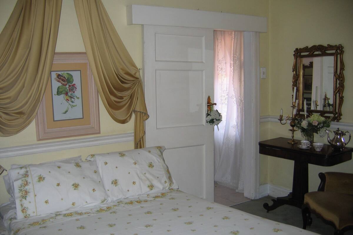 Un dormitorio con cama, mesita de noche con lámpara, espejo y puerta con cortinas.