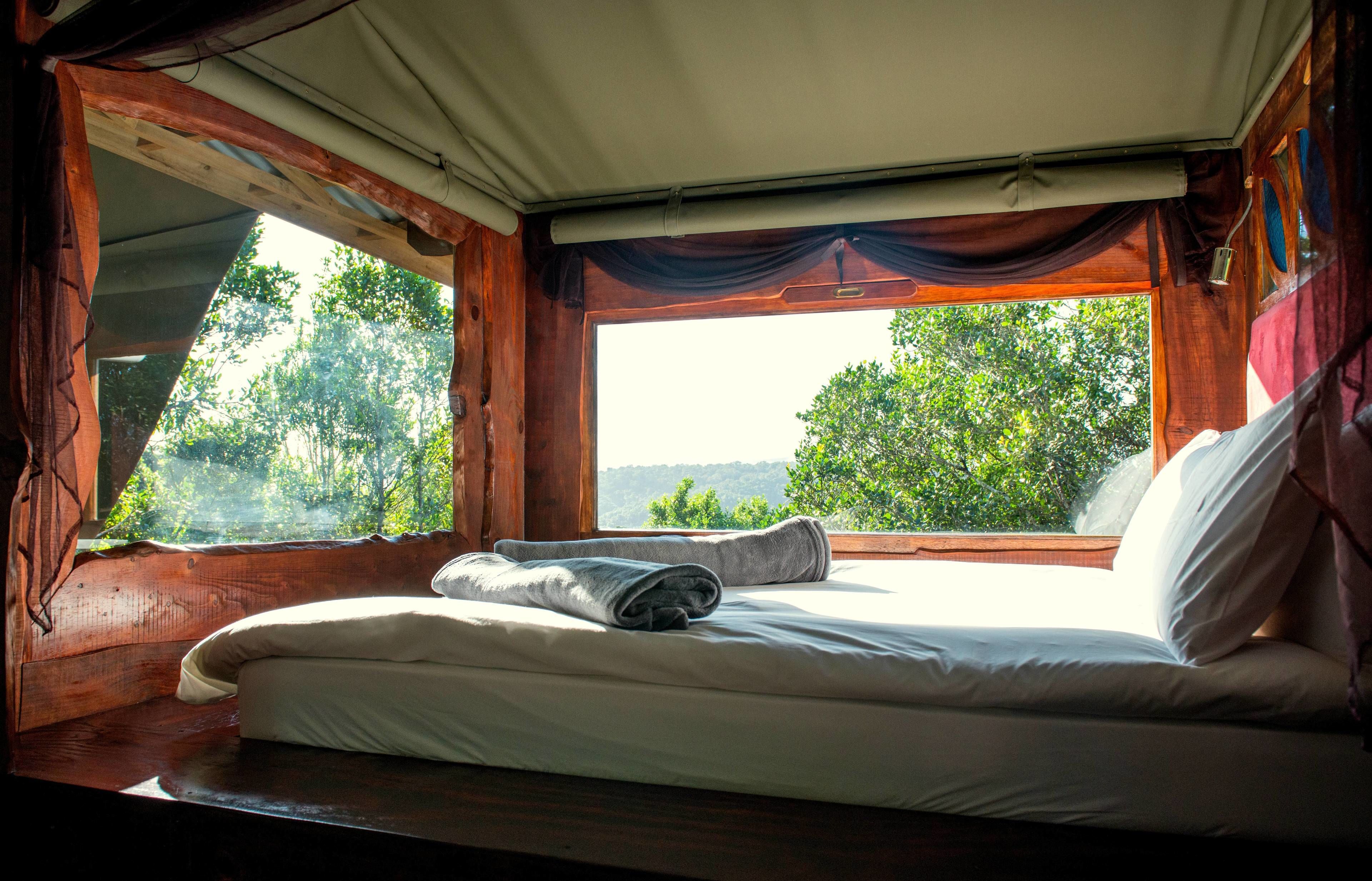 Cama com lençóis brancos e uma toalha enrolada, janela com vista para árvores e paredes de madeira.