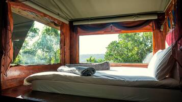 Cama com lençóis brancos e uma toalha enrolada, janela com vista para árvores e paredes de madeira.