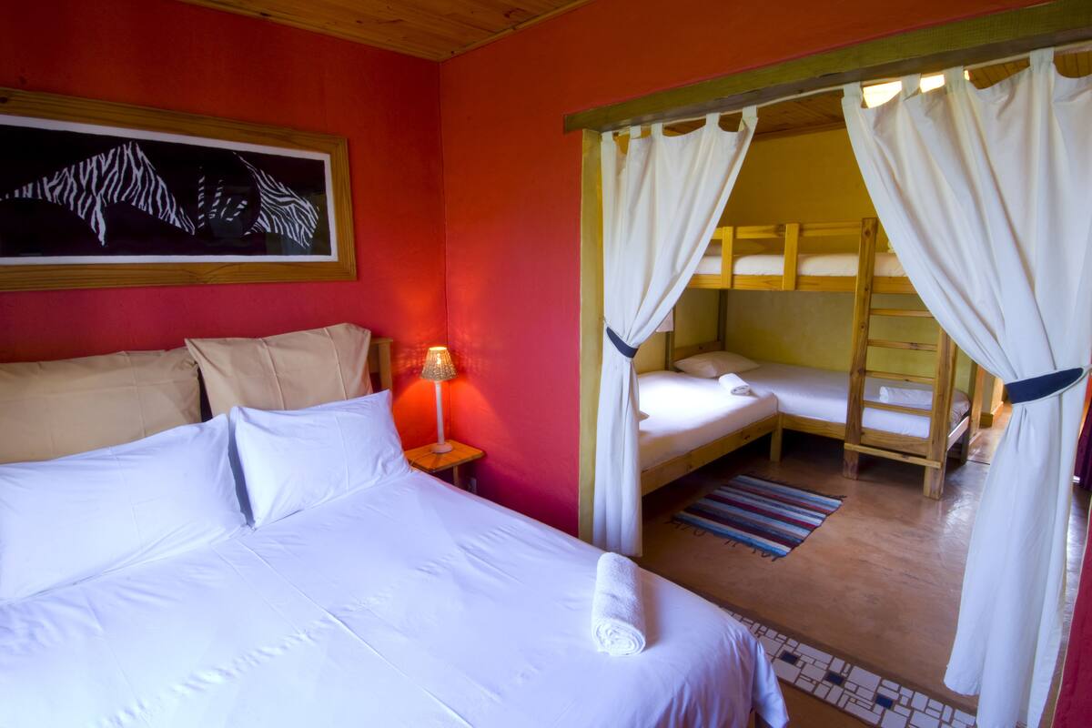 Una habitación con una cama grande, una cama más pequeña, una mesita de noche con lámpara y una litera.