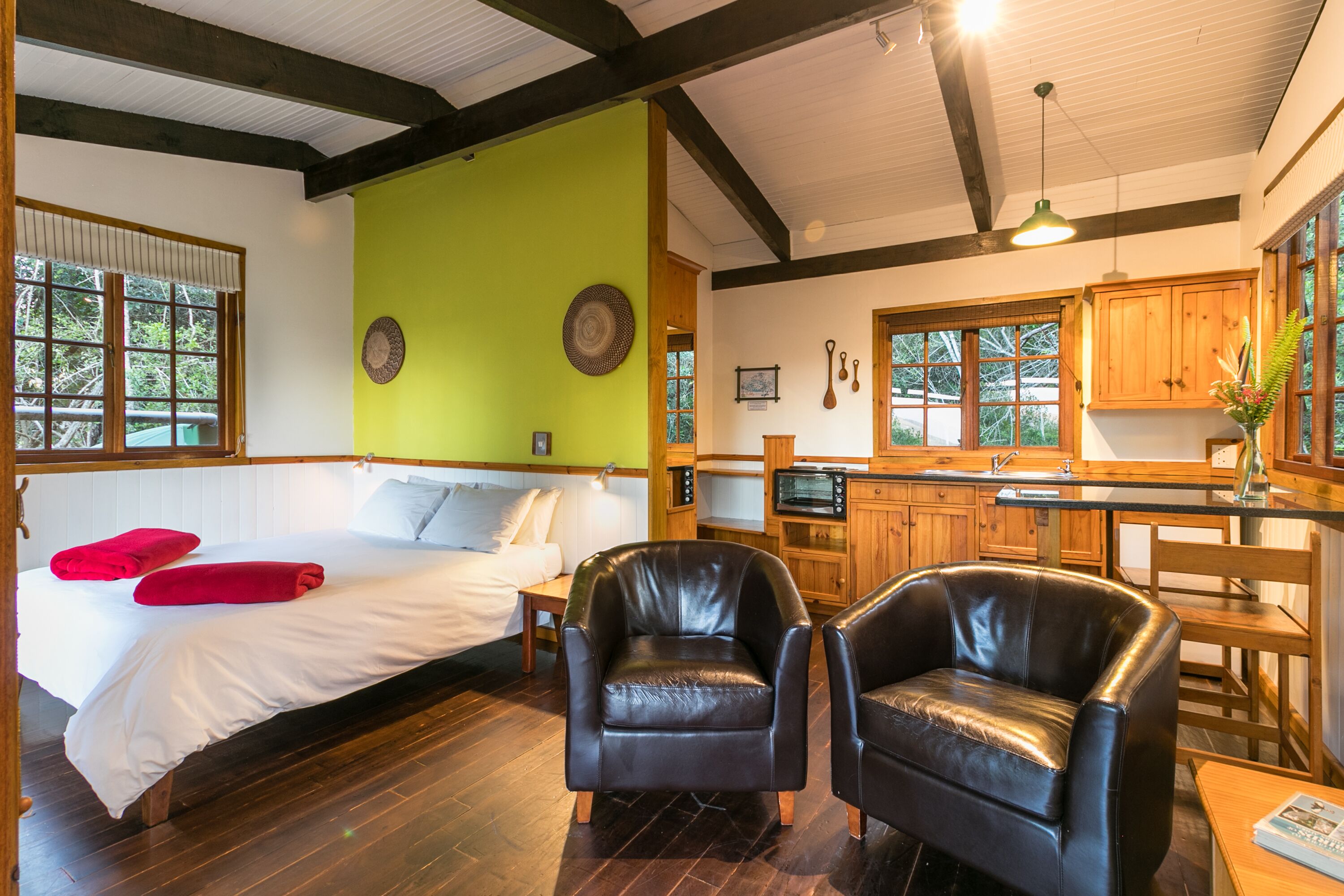 Habitación con una cama, dos sillones de cuero, una mesa de madera, una zona de cocina y una pared verde decorativa.