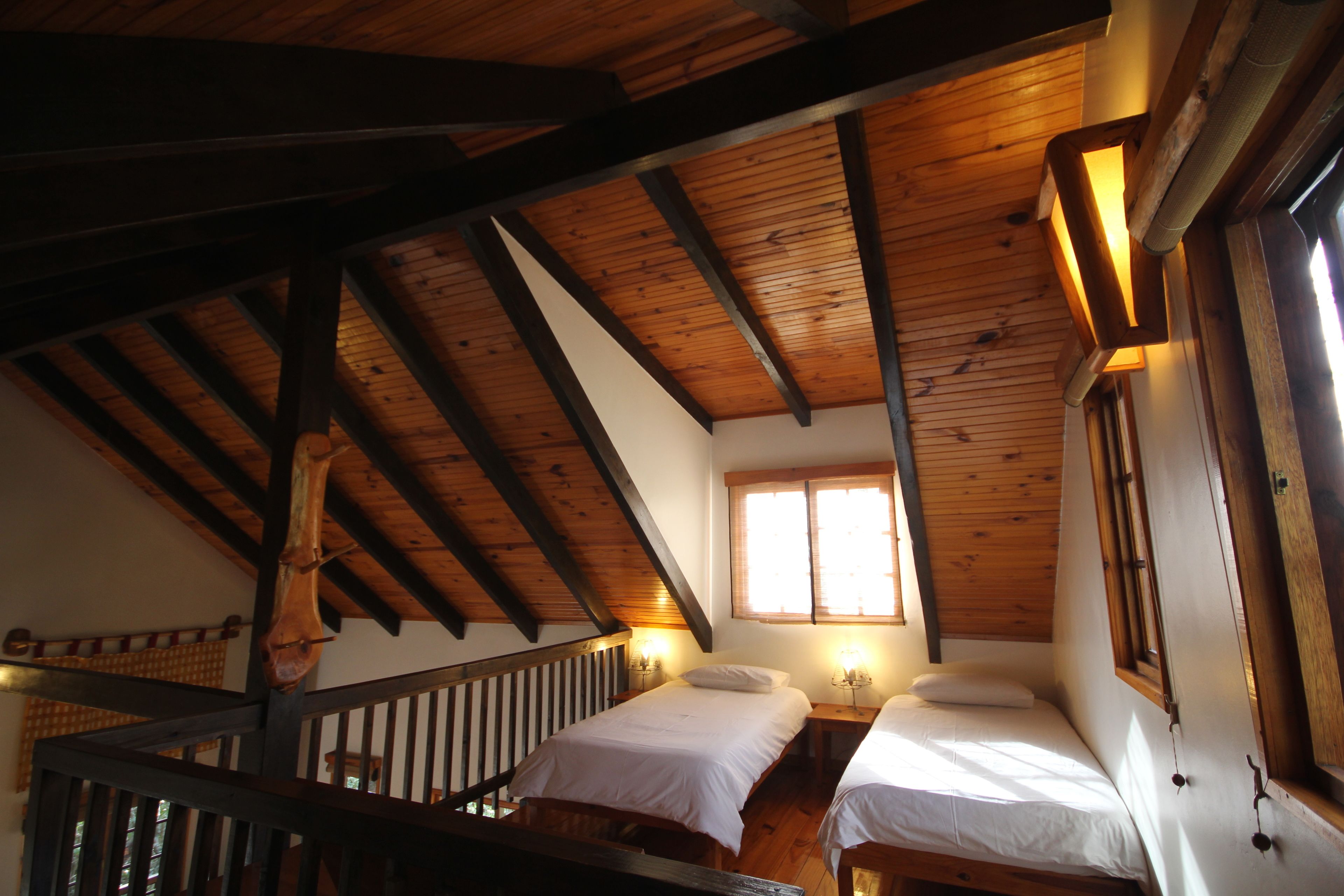 A cabin with two beds, wooden beams, a staircase, and a window with sunlight.