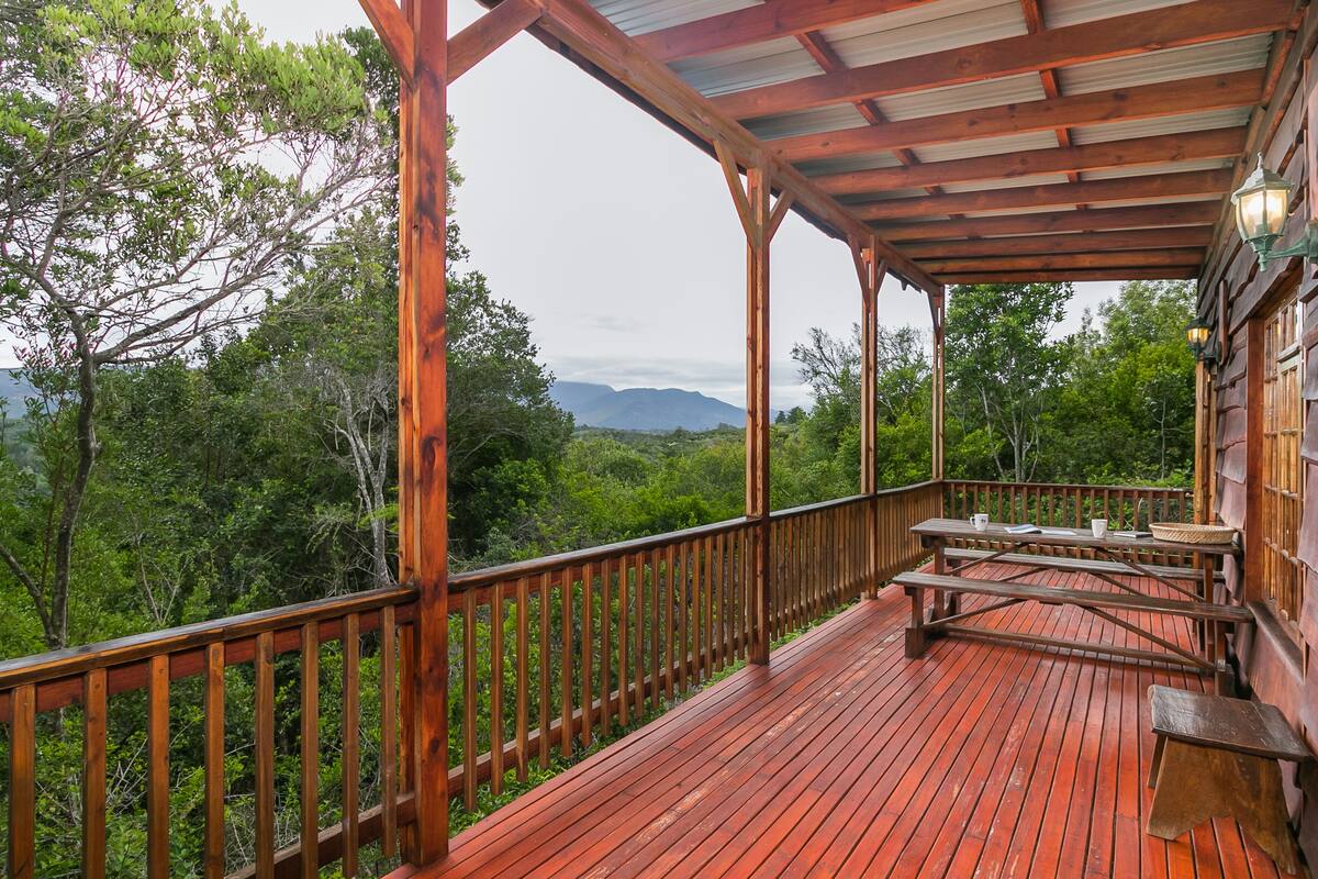 Una terraza de madera con mesas de picnic y bancos, con vistas a un área boscosa y montañas distantes.