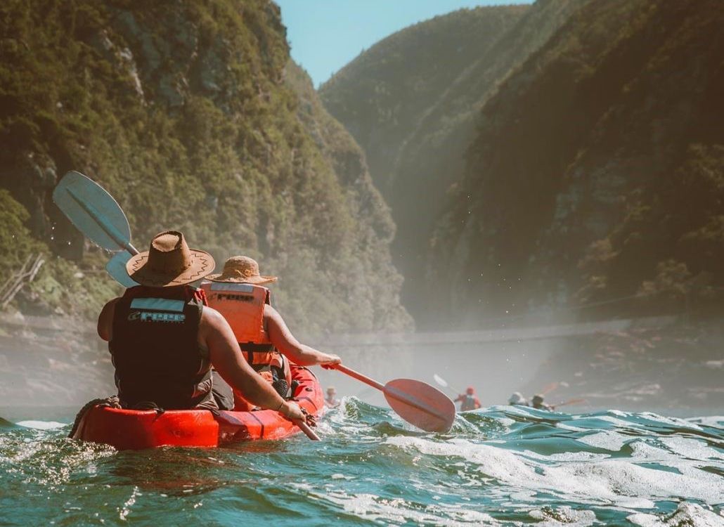 Duas pessoas em um bote inflável vermelho, com coletes salva-vidas, em meio a uma paisagem montanhosa.