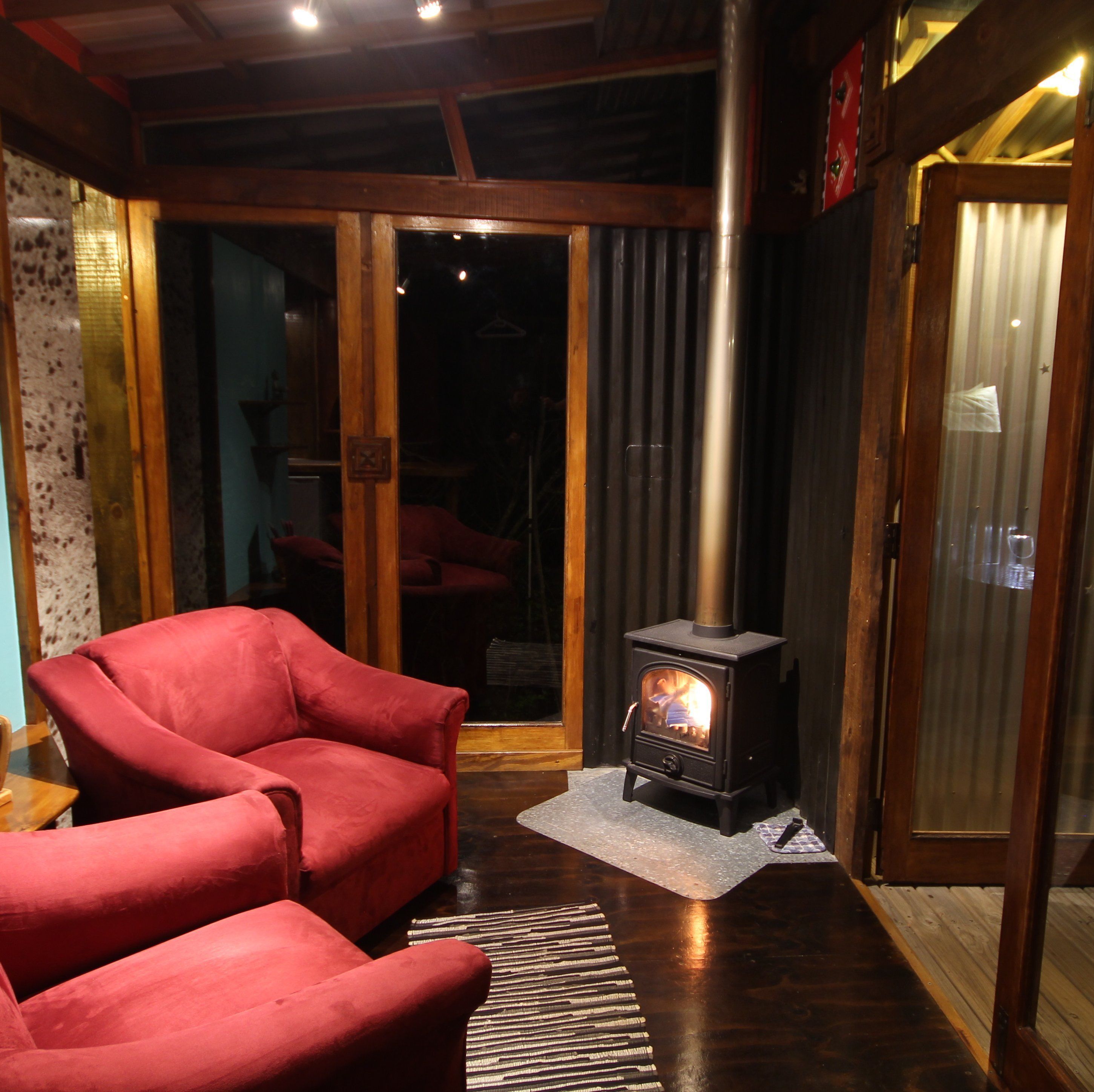 Una habitación acogedora con un sillón rojo, una estufa de leña y un ventanal corredero que da a un interior oscuro.
