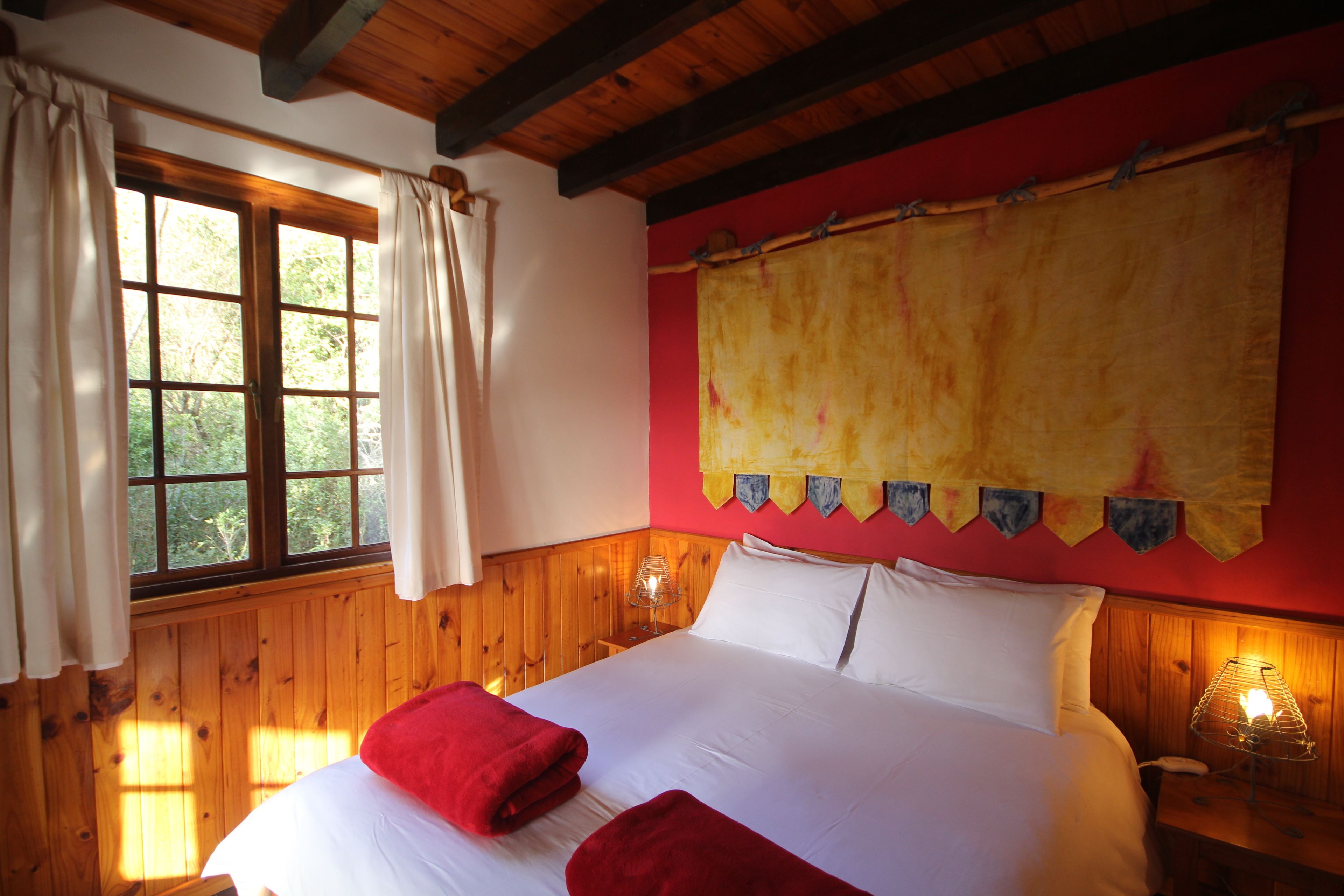 Una habitación con un revestimiento de madera, una cama con ropa blanca y toallas rojas, una ventana con cortinas y una lámpara fijada a la pared.