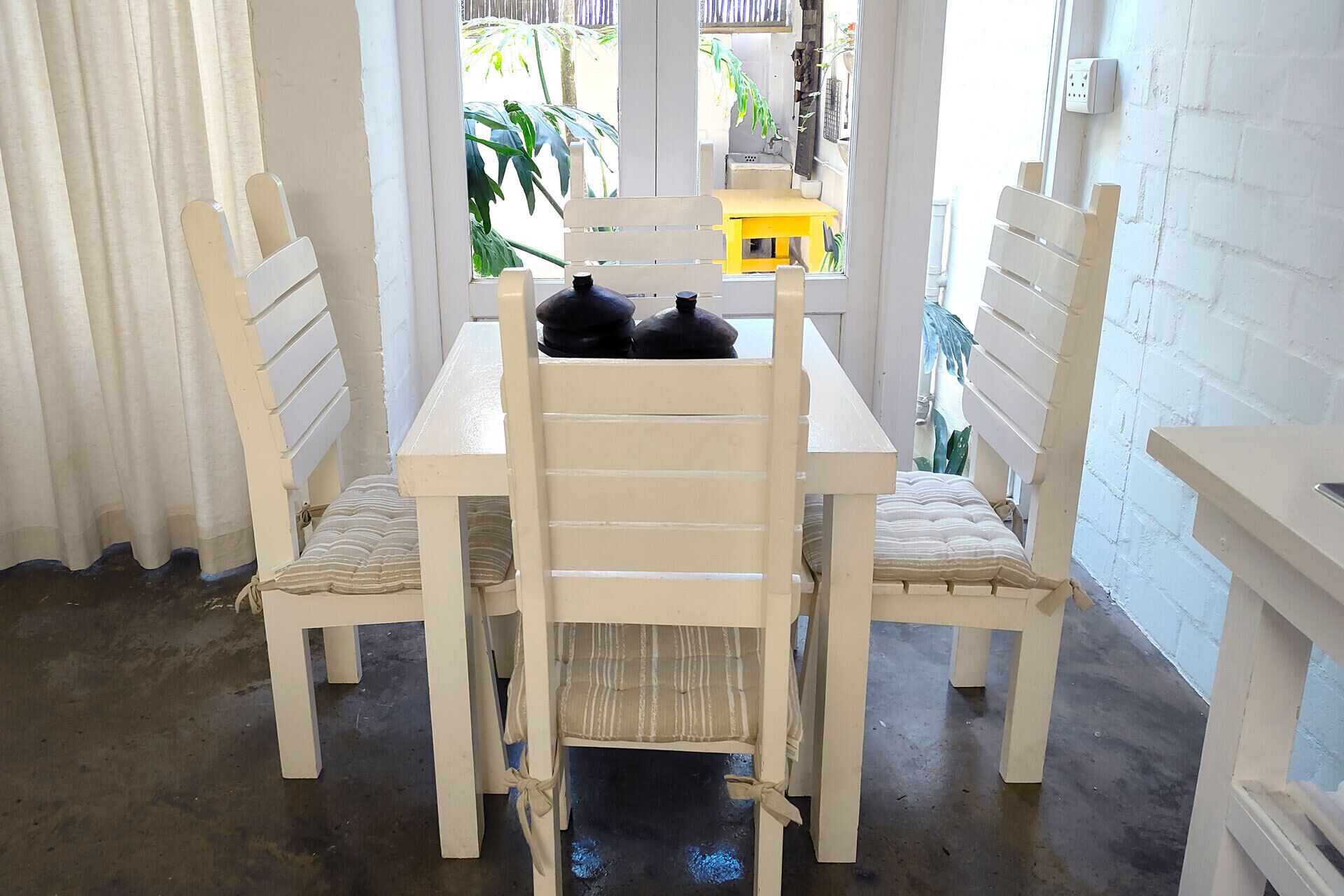 A white dining table with chairs, a yellow tablecloth, and a black object on the table.