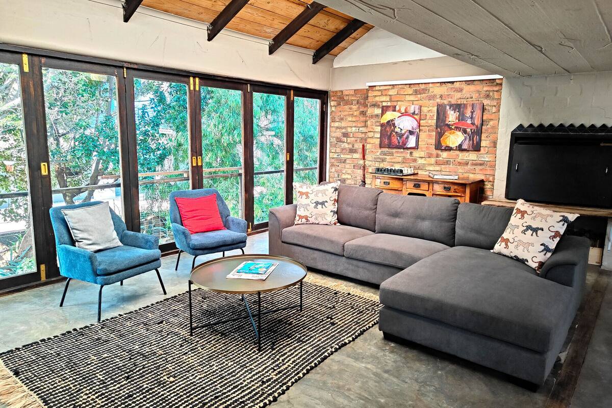 A modern living room with a grey sofa, blue armchair, and a coffee table. Large windows offer a view of greenery outside.