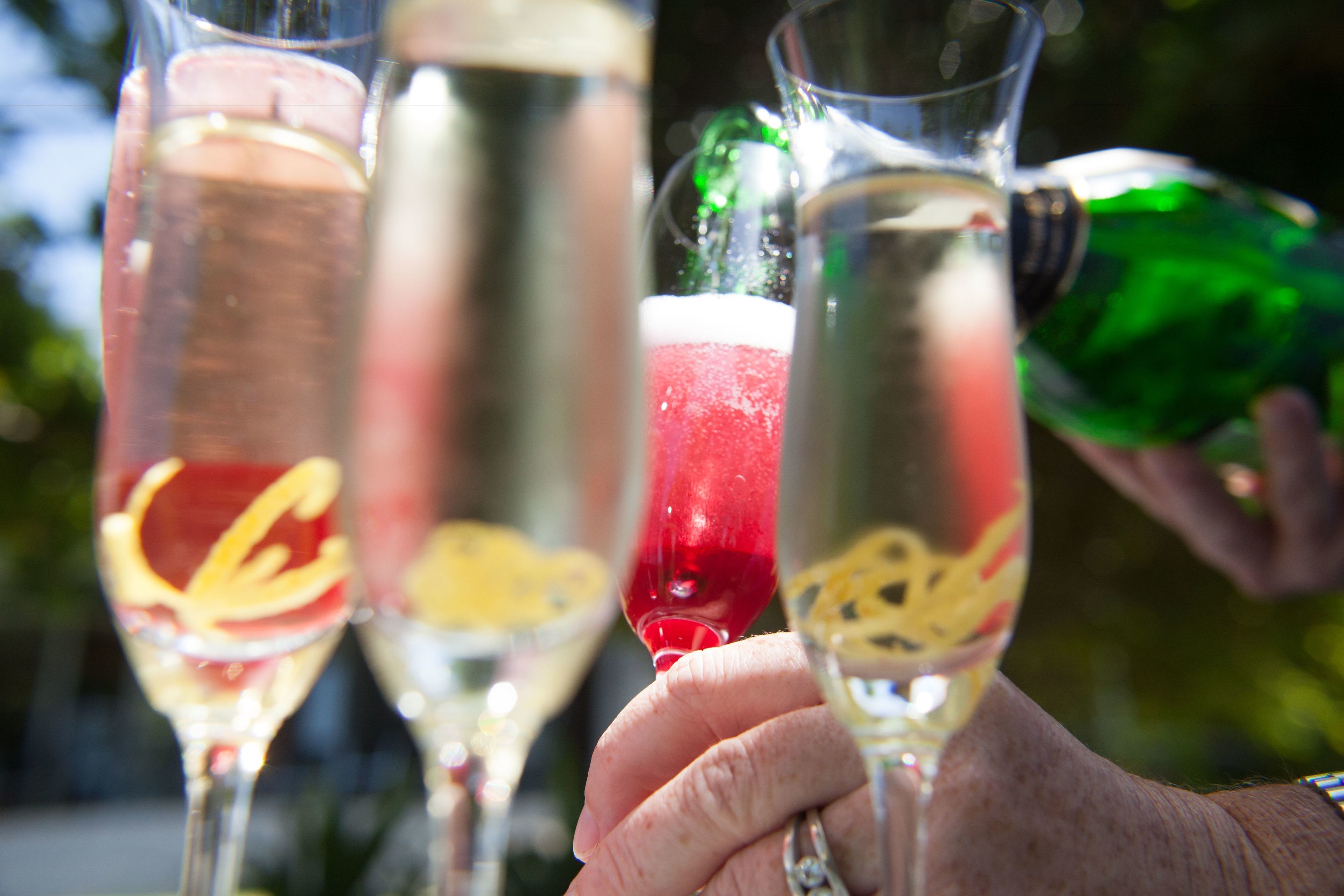 A close-up of hands holding champagne glasses with different colored liquids, and a green bottle being poured into one of the glasses.