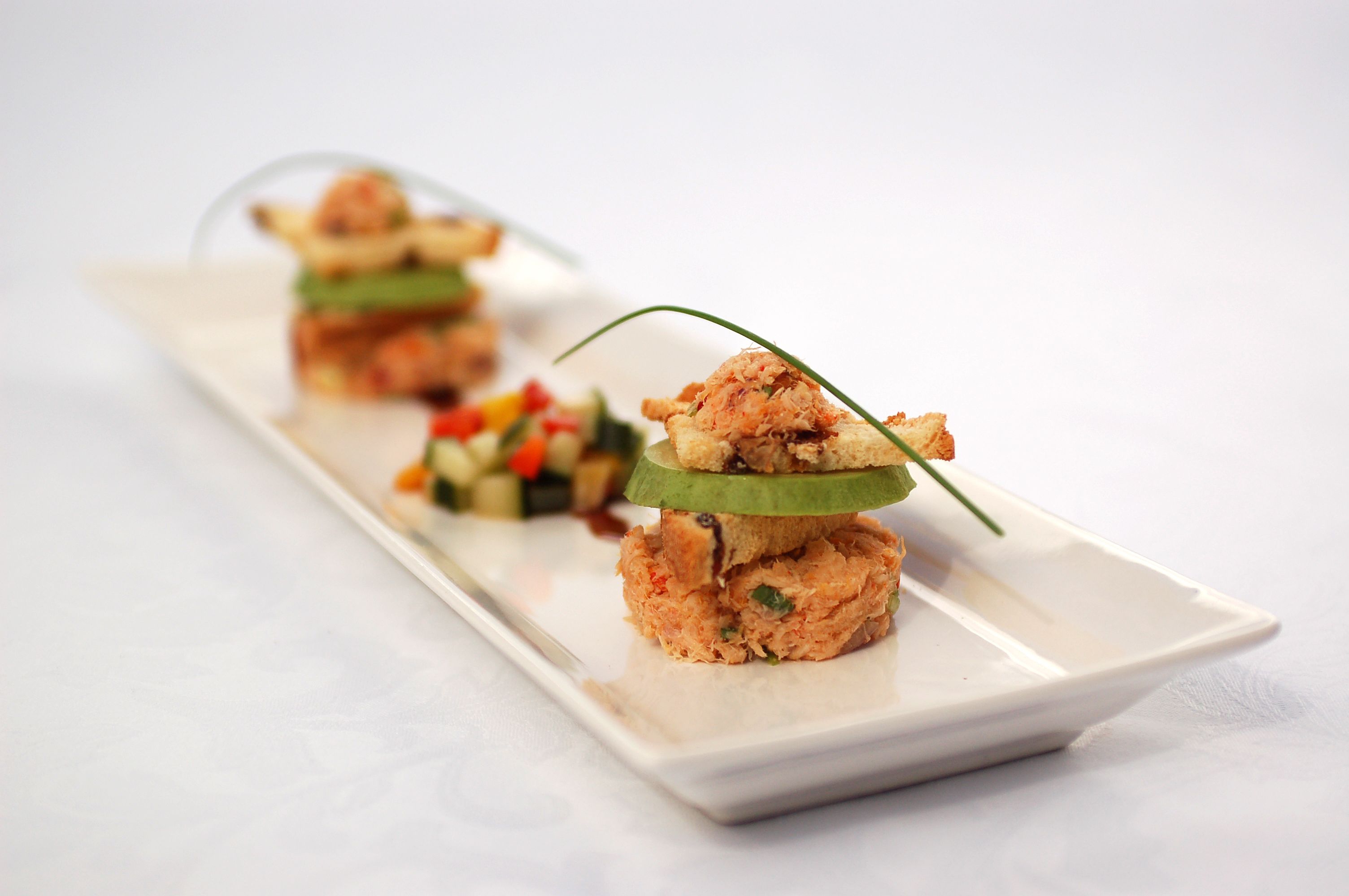 A white rectangular plate with three stacked food items, each with a slice of cucumber on top.