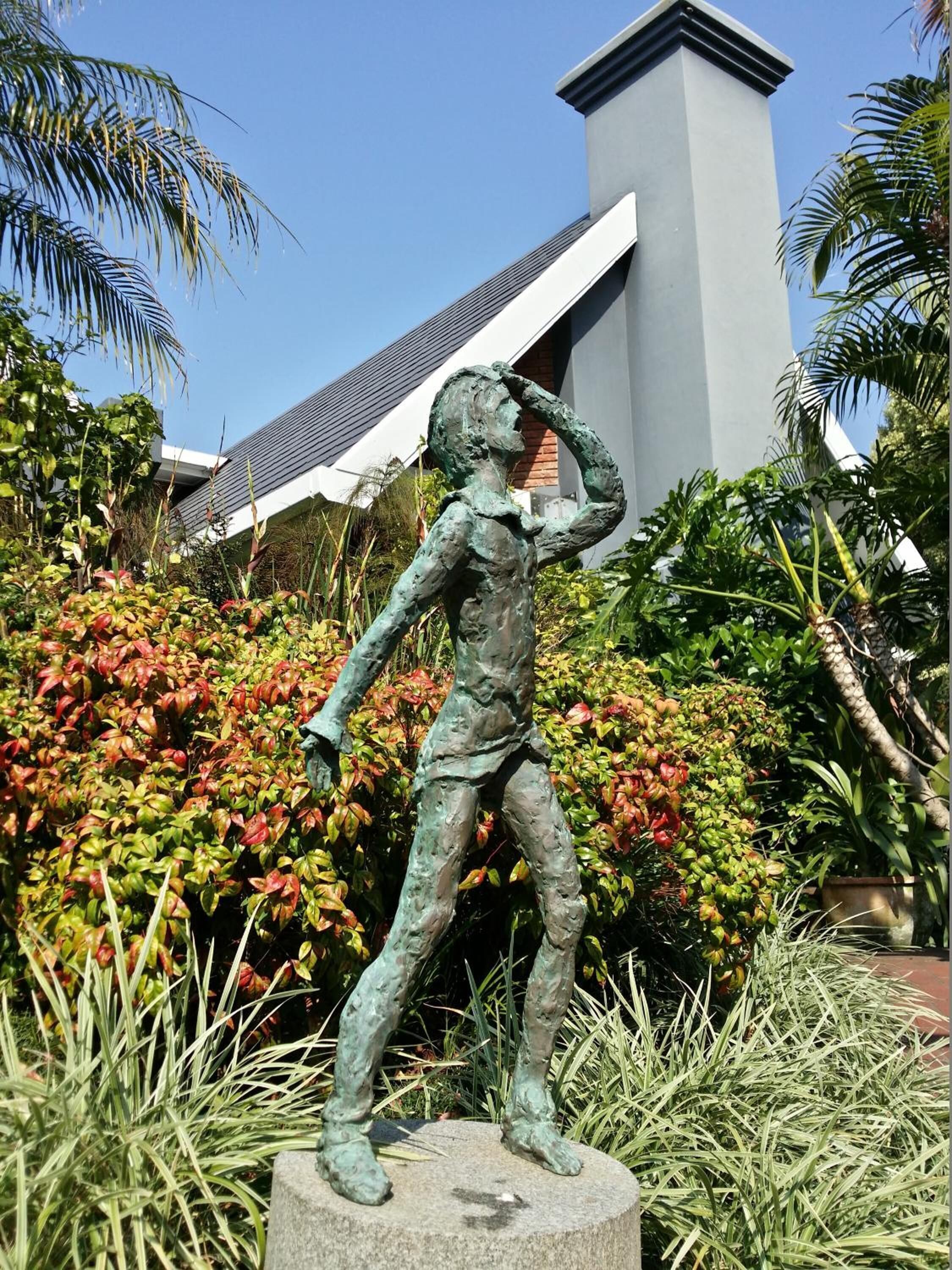 Une statue de bronze représentant un homme dans une pose dynamique, placée sur un piédestal en pierre dans un jardin où poussent des plantes tropicales, avec un bâtiment moderne en arrière-plan.