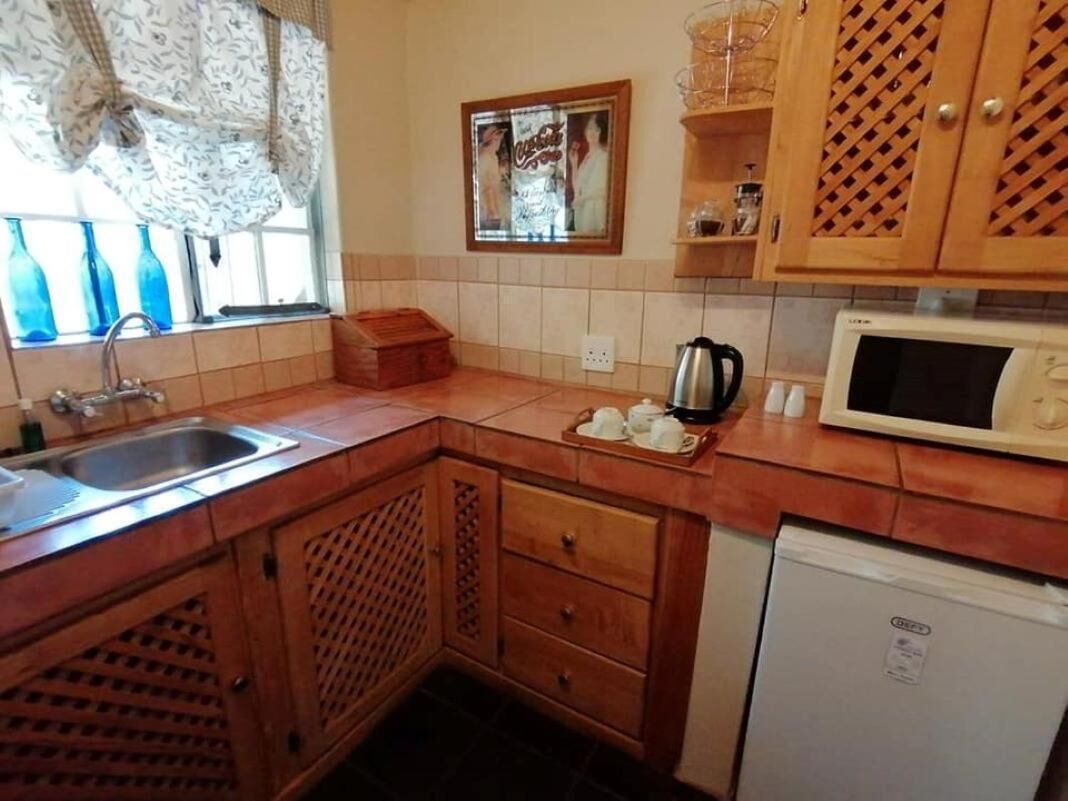 Cucina compatta con lavello, microonde e piccolo frigorifero. Armadietti in legno con decoro a reticolo, e finestra con tende a fantasia.