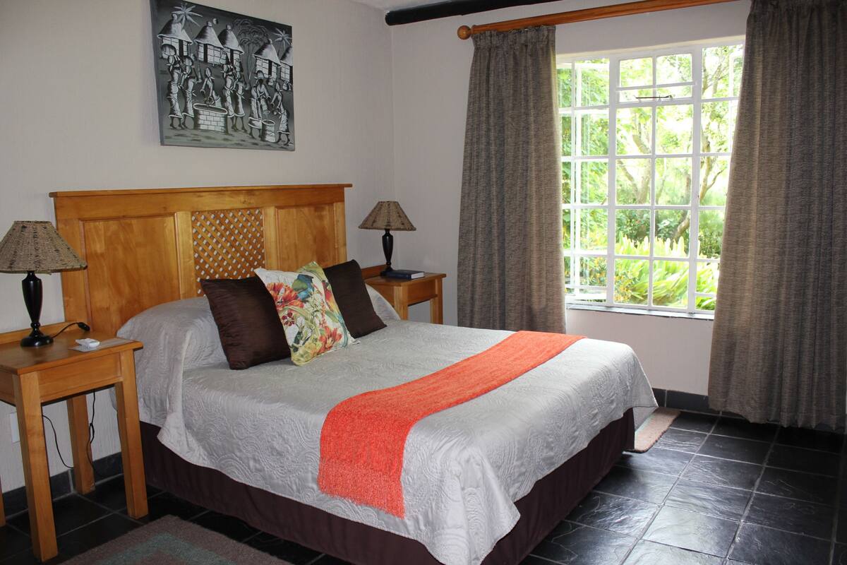 A bedroom with a large bed, two bedside tables with lamps, a window with curtains, and a wall-mounted painting.