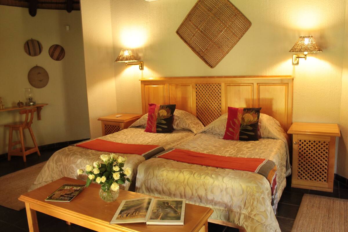 A double room with two beds, a wooden headboard, bedside tables, a nightstand, a small table with a vase of flowers, and a wall-mounted lamp.