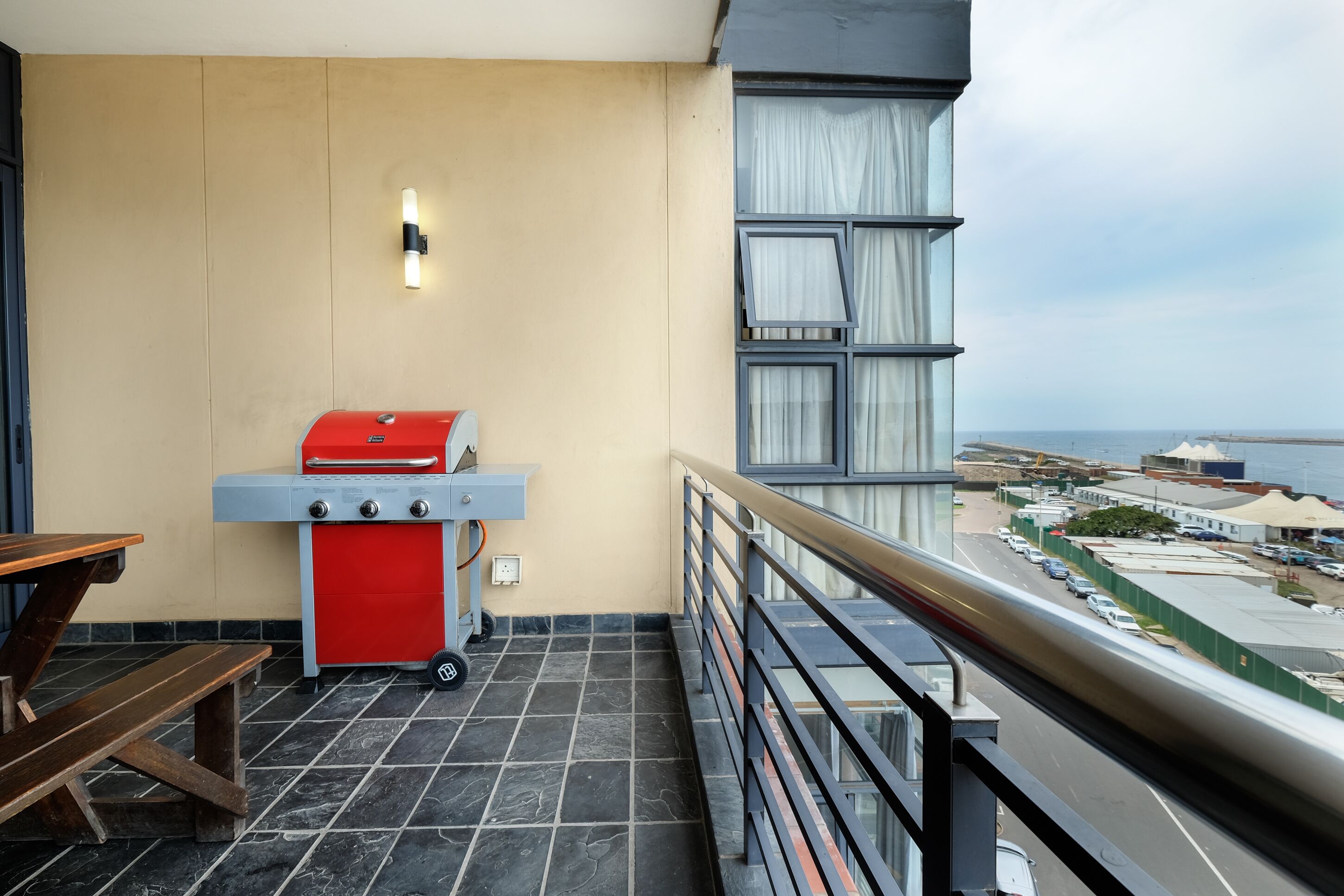 A balcony with a red grill, a table, and a view of the sea and buildings.