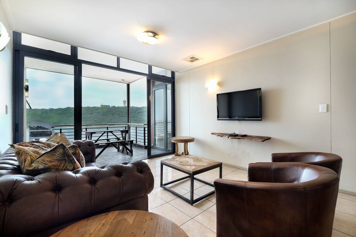 A modern living room with a leather sofa, a coffee table, and leather armchairs. A flat-screen TV is mounted on the wall, and there is a balcony with a table and chairs.