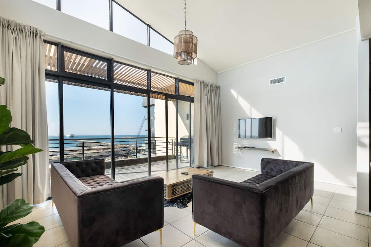 A modern living room with large windows, a flat-screen TV, and two dark sofas.