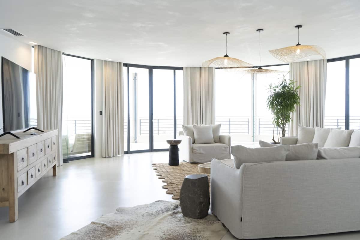 A modern living room with a sofa, a coffee table, and a TV on a wooden cabinet. There are large windows with curtains and a plant in the corner.