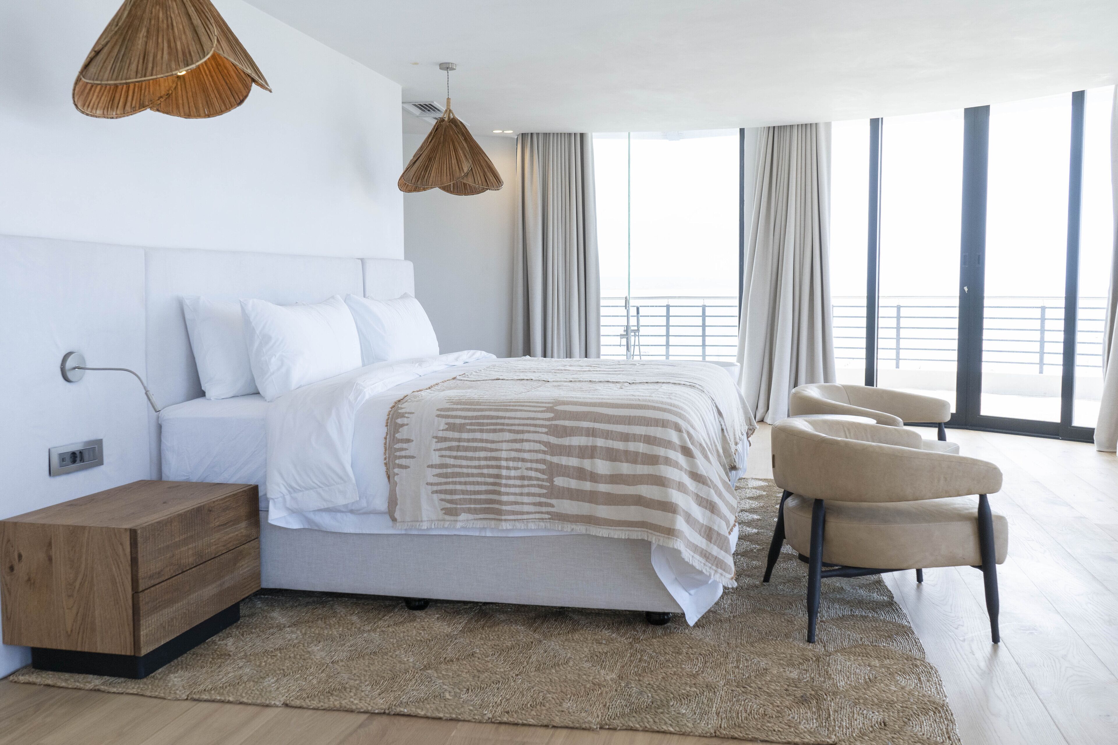 A modern bedroom with a large bed, a wooden nightstand, a chair, and a view of the balcony.