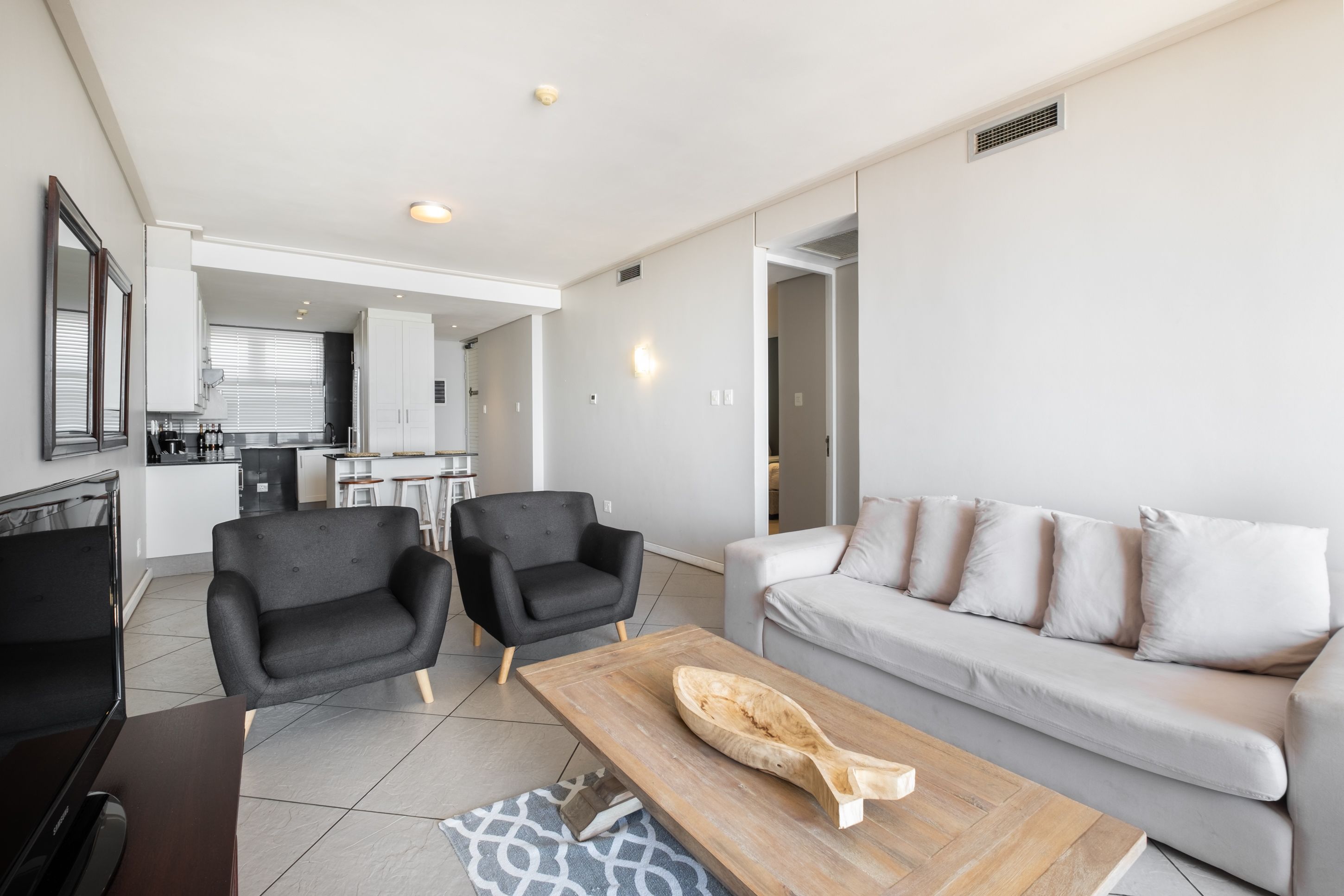 A modern living room with a grey sofa, two black armchairs, a wooden coffee table, and a kitchen in the background.