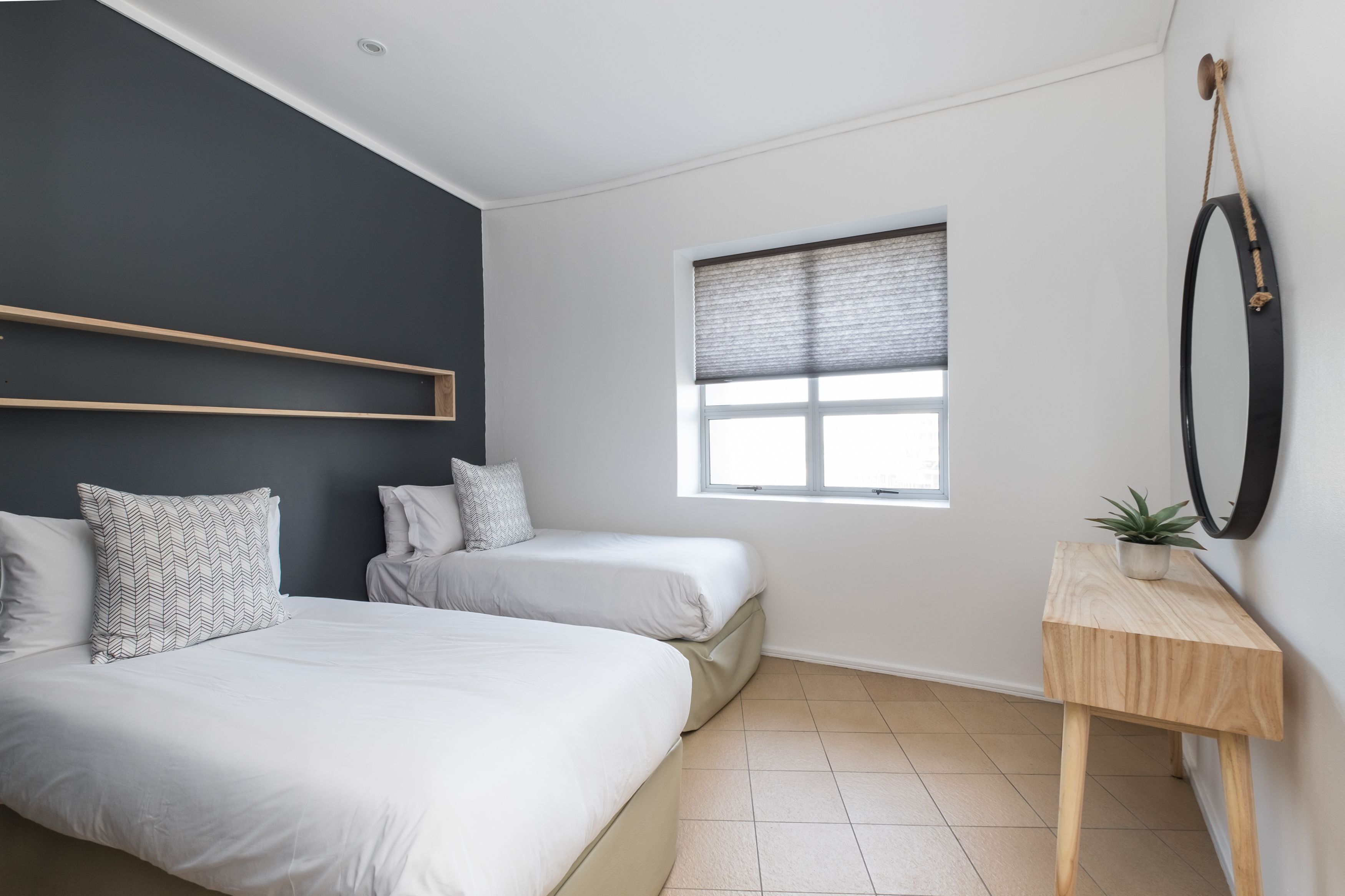 A modern bedroom with two beds, a window with blinds, a wooden bedside table with a plant, and a dark accent wall.