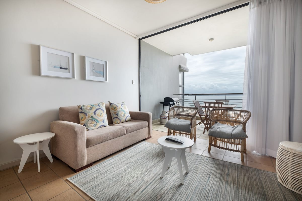 A modern living room with a sofa, a small table, and wicker chairs. There are two framed pictures on the wall, and a view of the sea through a large window.