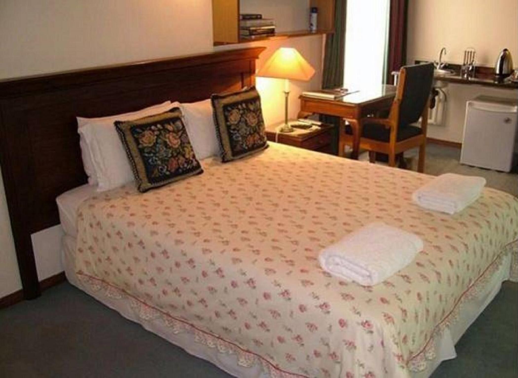 A neatly made bed with floral bedding, a bedside lamp, a small desk with a chair, and a refrigerator in a hotel room.