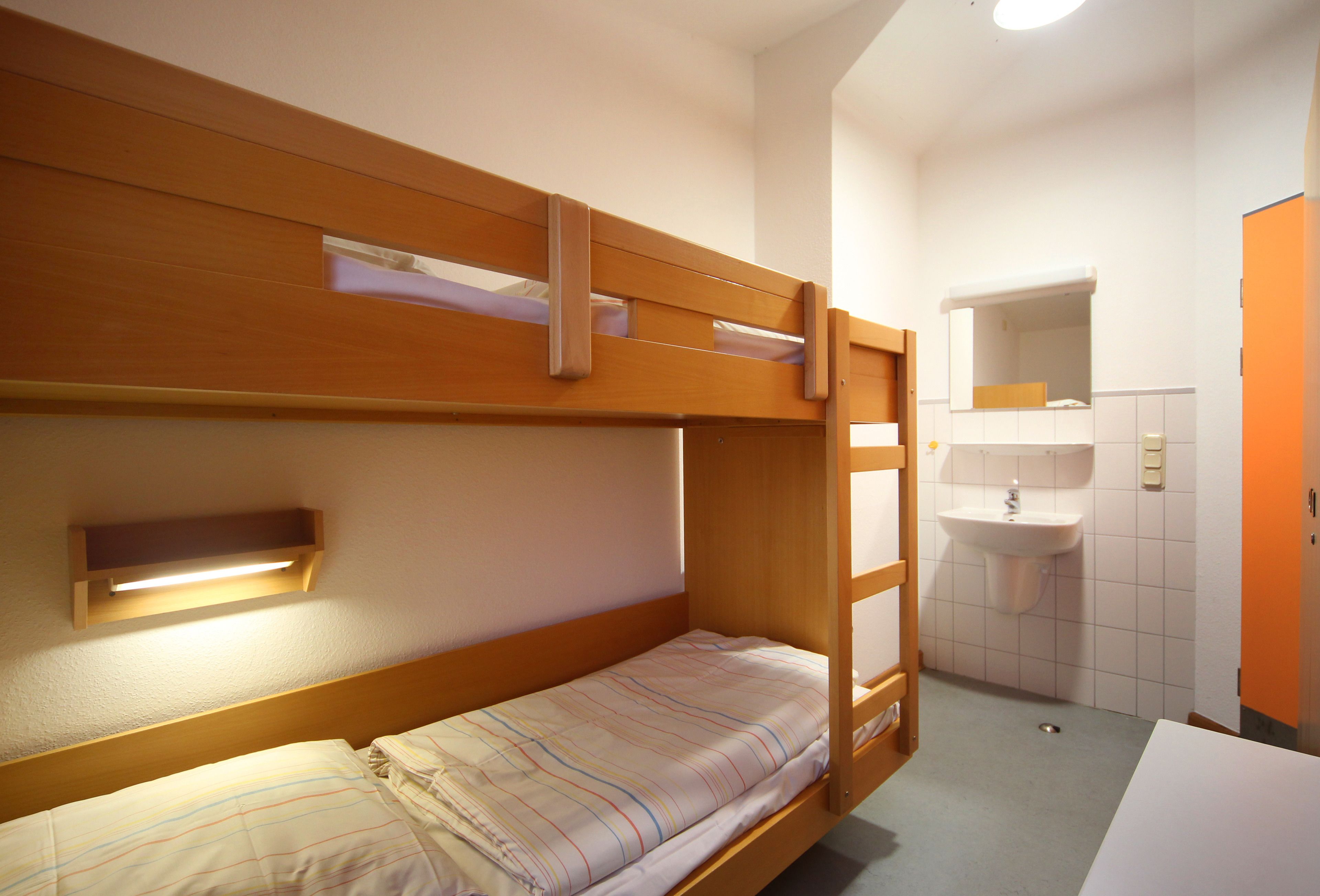 Une chambre avec un lit superposé, une table de chevet et une petite étagère. La salle de bain est équipée d’un lavabo et d’un miroir.