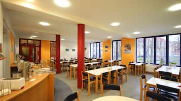 Eine Cafeteria mit mehreren Tischen, Stühlen, einer Speisenausgabe und großen Fenstern, die für natürliches Licht sorgen.