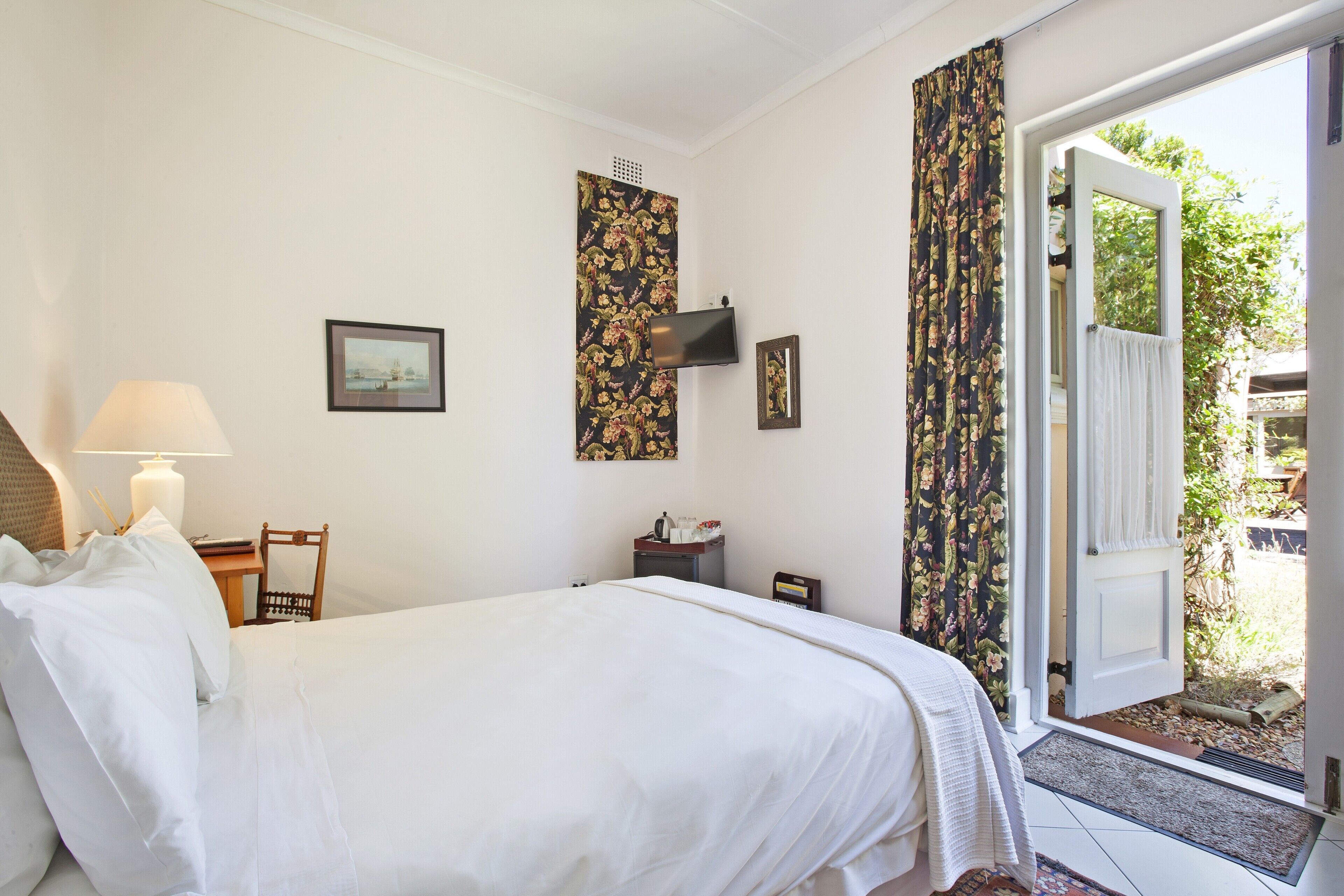 Habitación de hotel con una cama, un escritorio con lámpara, una silla, un televisor y una puerta que da a un área exterior.