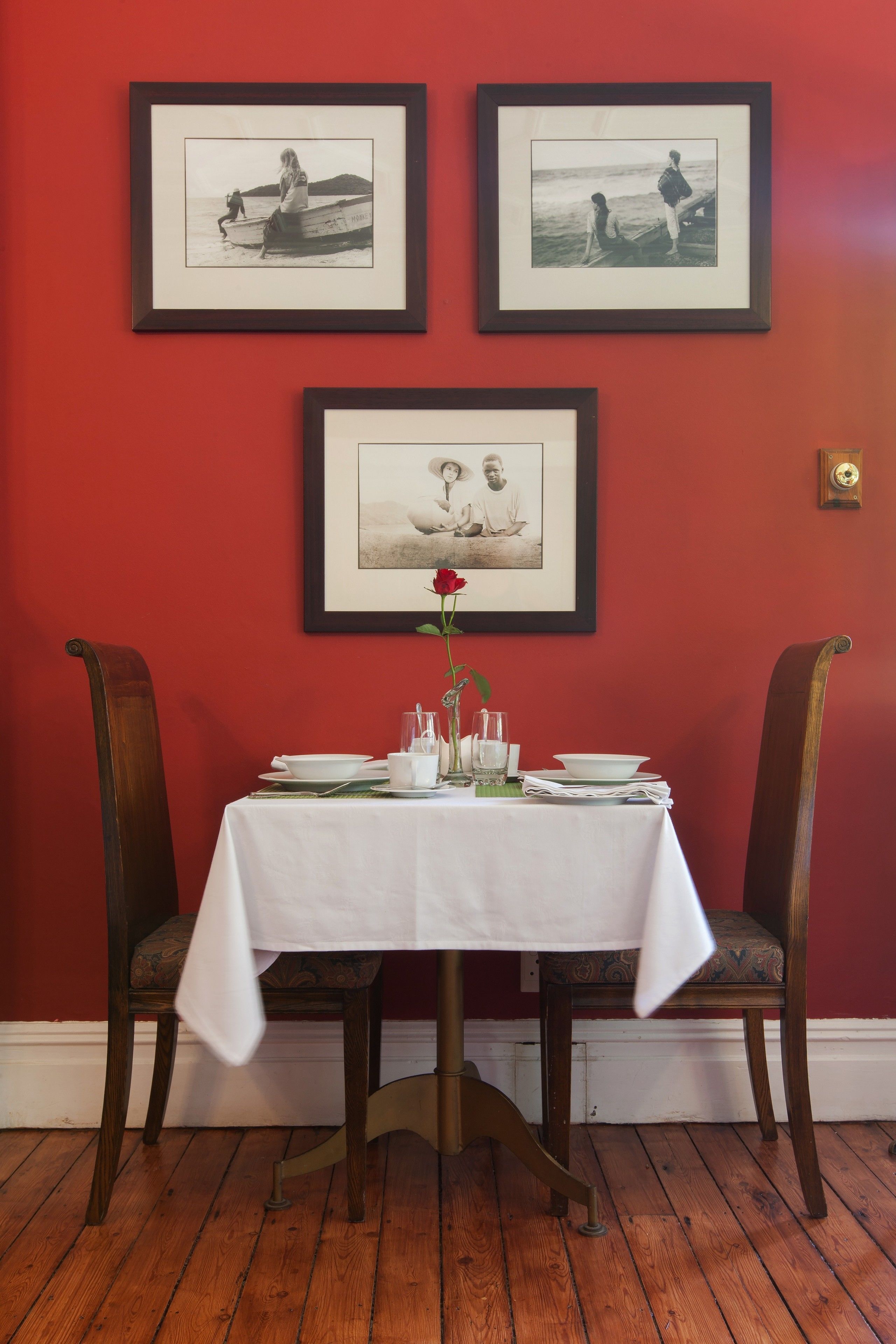 A dining table set with plates, glasses, and a vase with a single flower, flanked by two chairs, against a red wall with three framed pictures above.