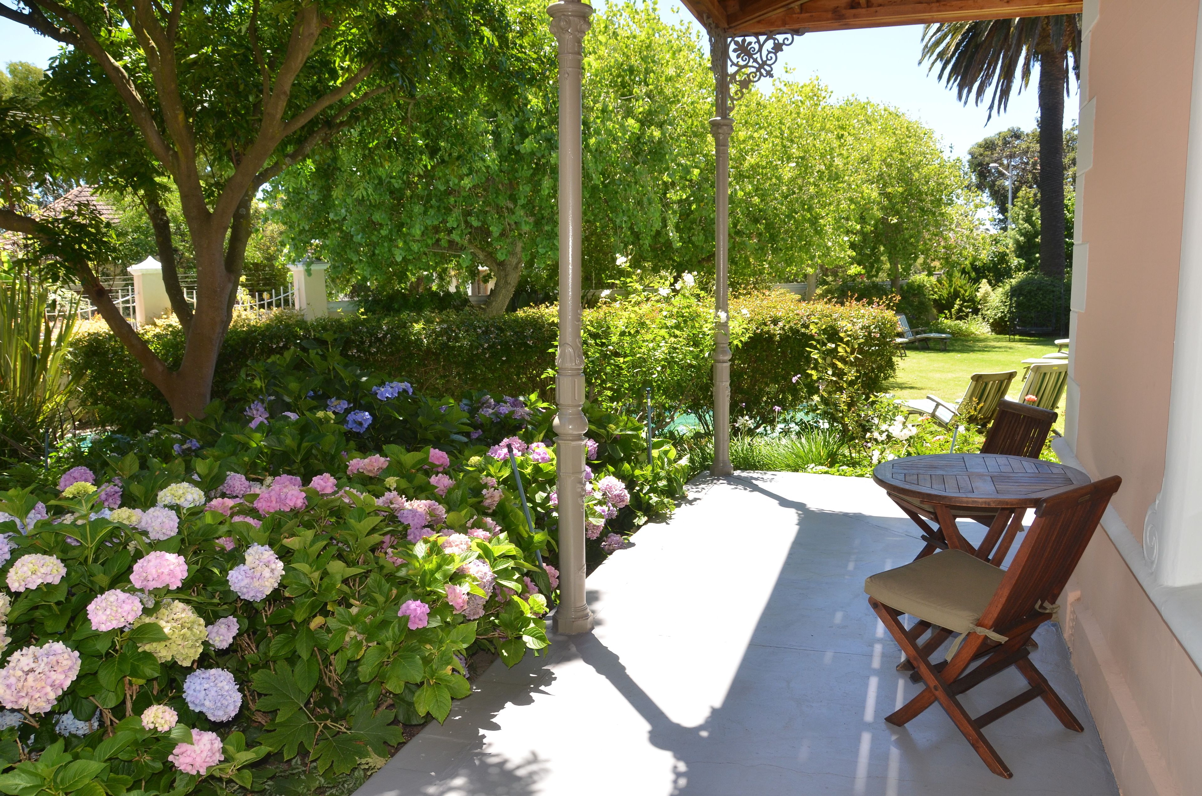 A covered patio with a table and chairs, surrounded by lush greenery and flowering plants.