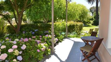 A covered patio with a table and chairs, surrounded by lush greenery and flowering plants.