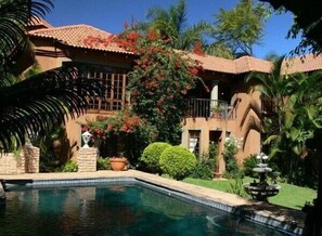 A pool with a fountain, surrounded by lush greenery and a building with a tiled roof.