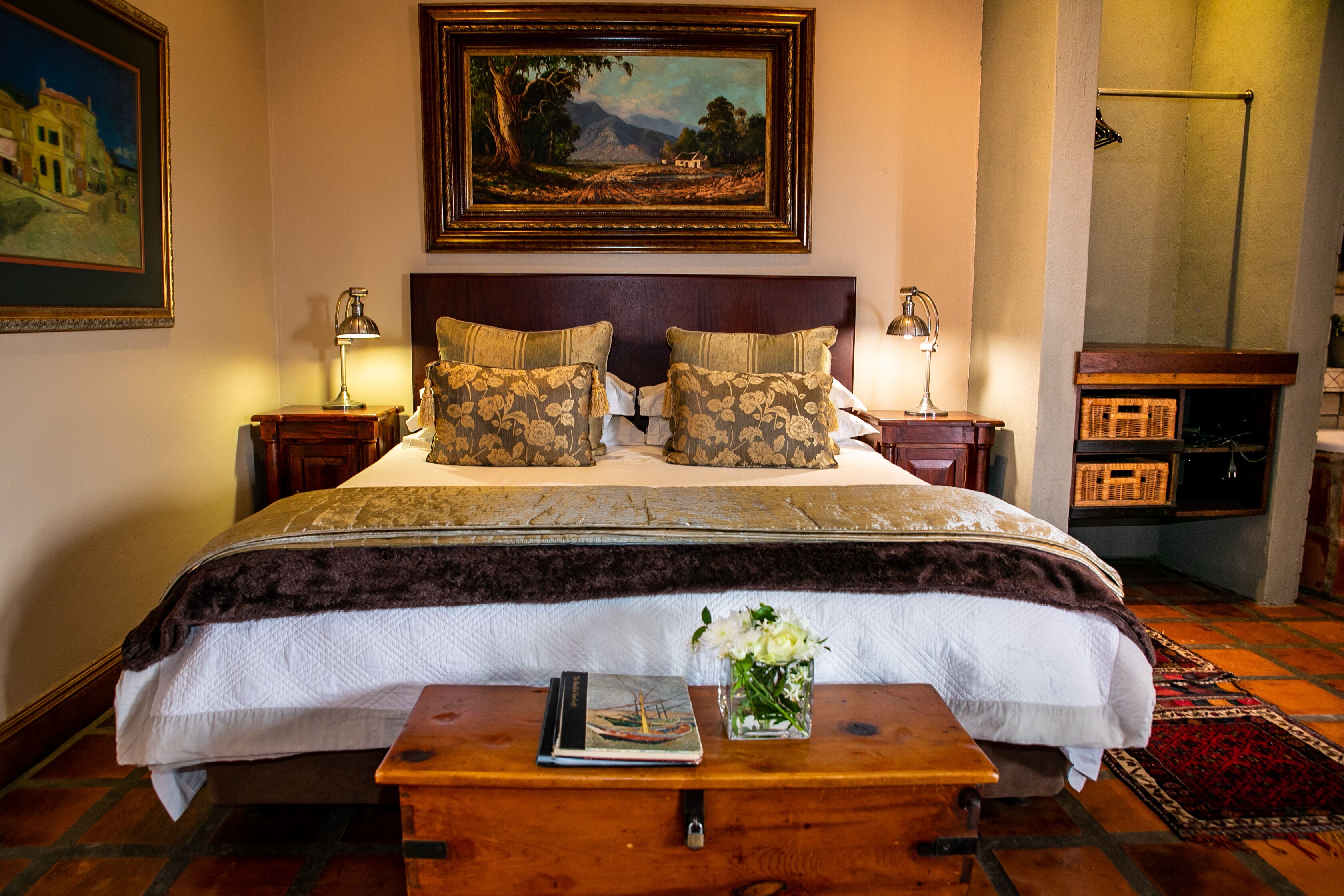 A neatly arranged bedroom with a large bed, two bedside tables with lamps, a wooden nightstand, a small table with a book and flowers, and a painting on the wall.