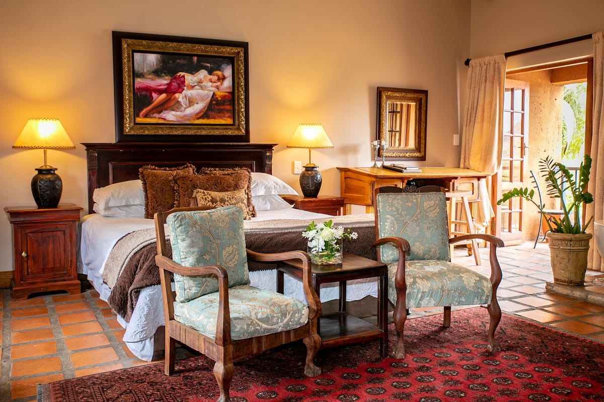 A bedroom with a bed, two lamps, a desk, two chairs, a painting, and a potted plant.