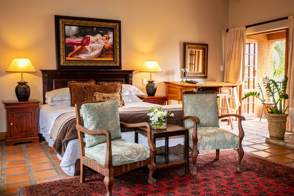 A bedroom with a bed, two lamps, a desk, two chairs, a painting, and a potted plant.