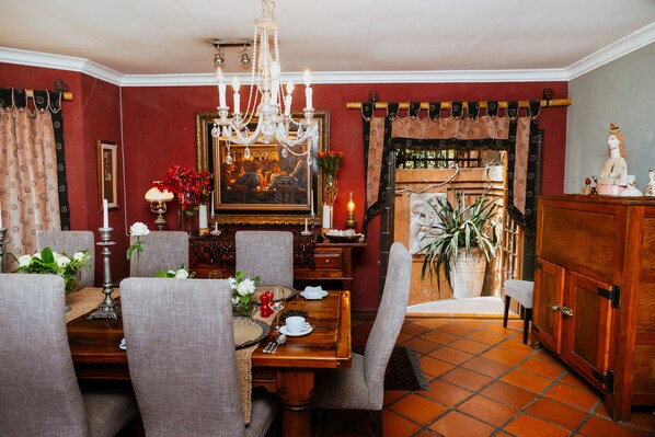 Comedor con mesa de madera preparada para cuatro personas, rodeada de sillas tapizadas. Un candelabro cuelga del techo, y hay un cuadro enmarcado en la pared.